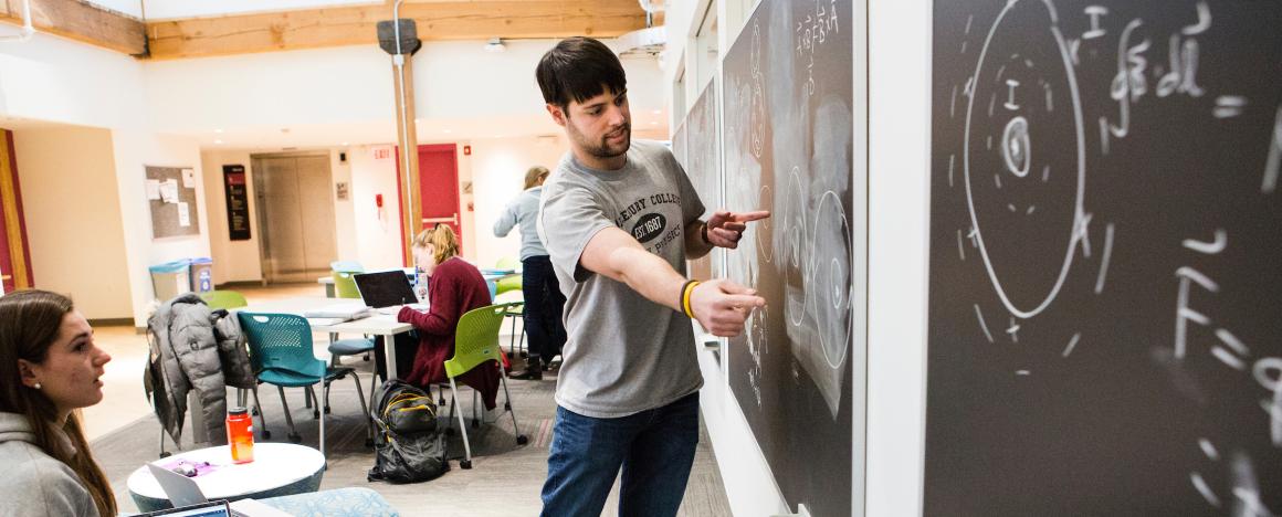 A student discusses an equation on a chalkboard with another student in a large, brightly lit room